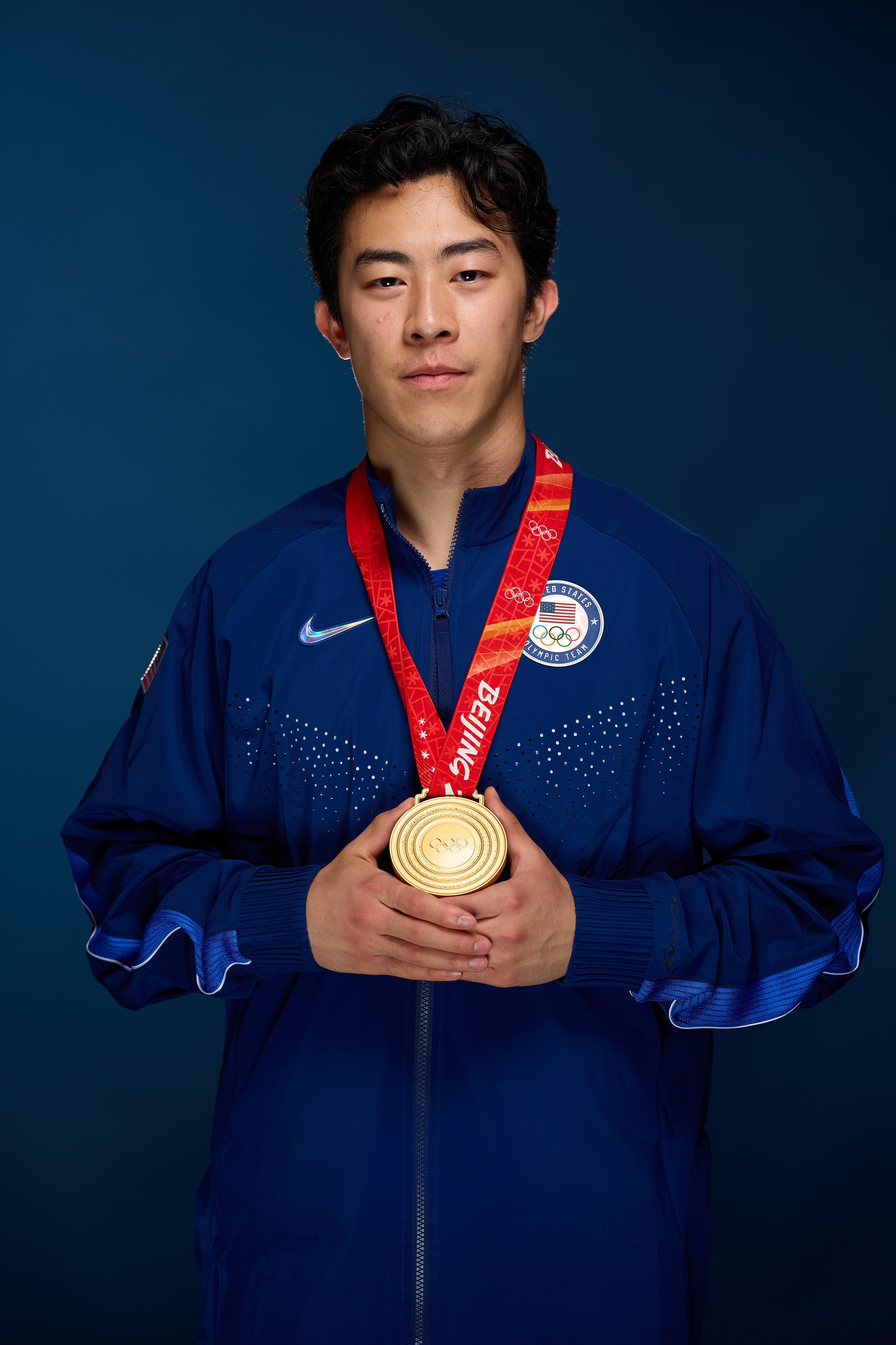 Athlete holds a medal, wearing a sporty overgarment   with a logo. Dominant expression, posed against a plain background