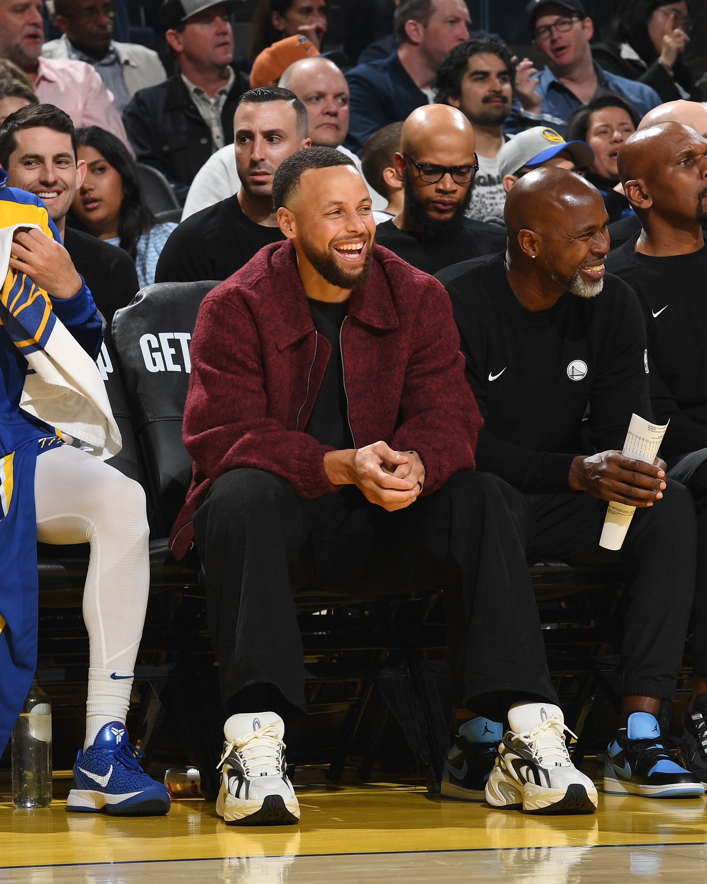 A antheral   successful  a maroon overgarment   and sneakers sits smiling courtside among others astatine  a hoops  game