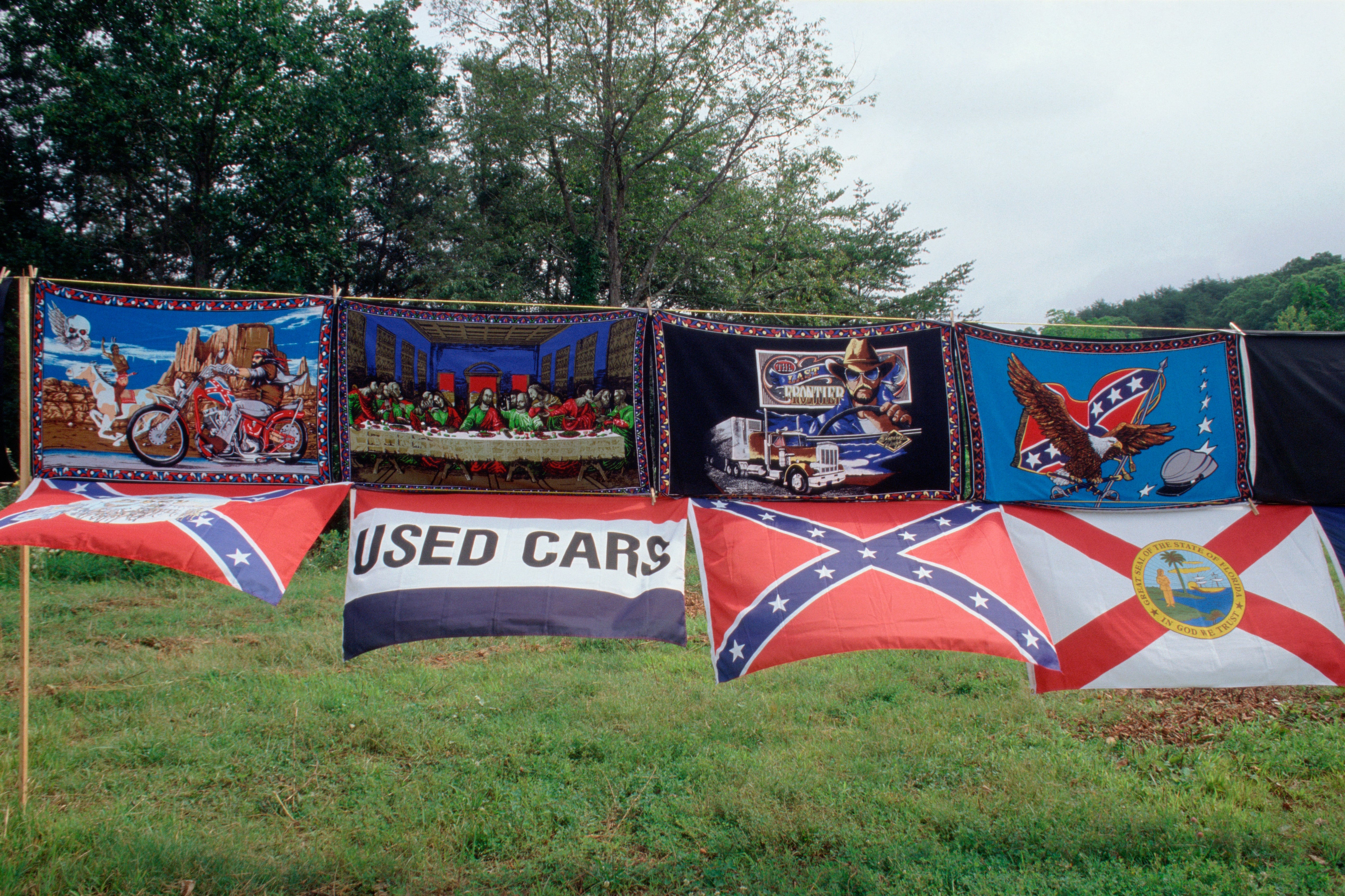 Various flags and blankets bent  connected  a enactment     outdoors, including respective  with Confederate imagery and 1  with a utilized  cars sign