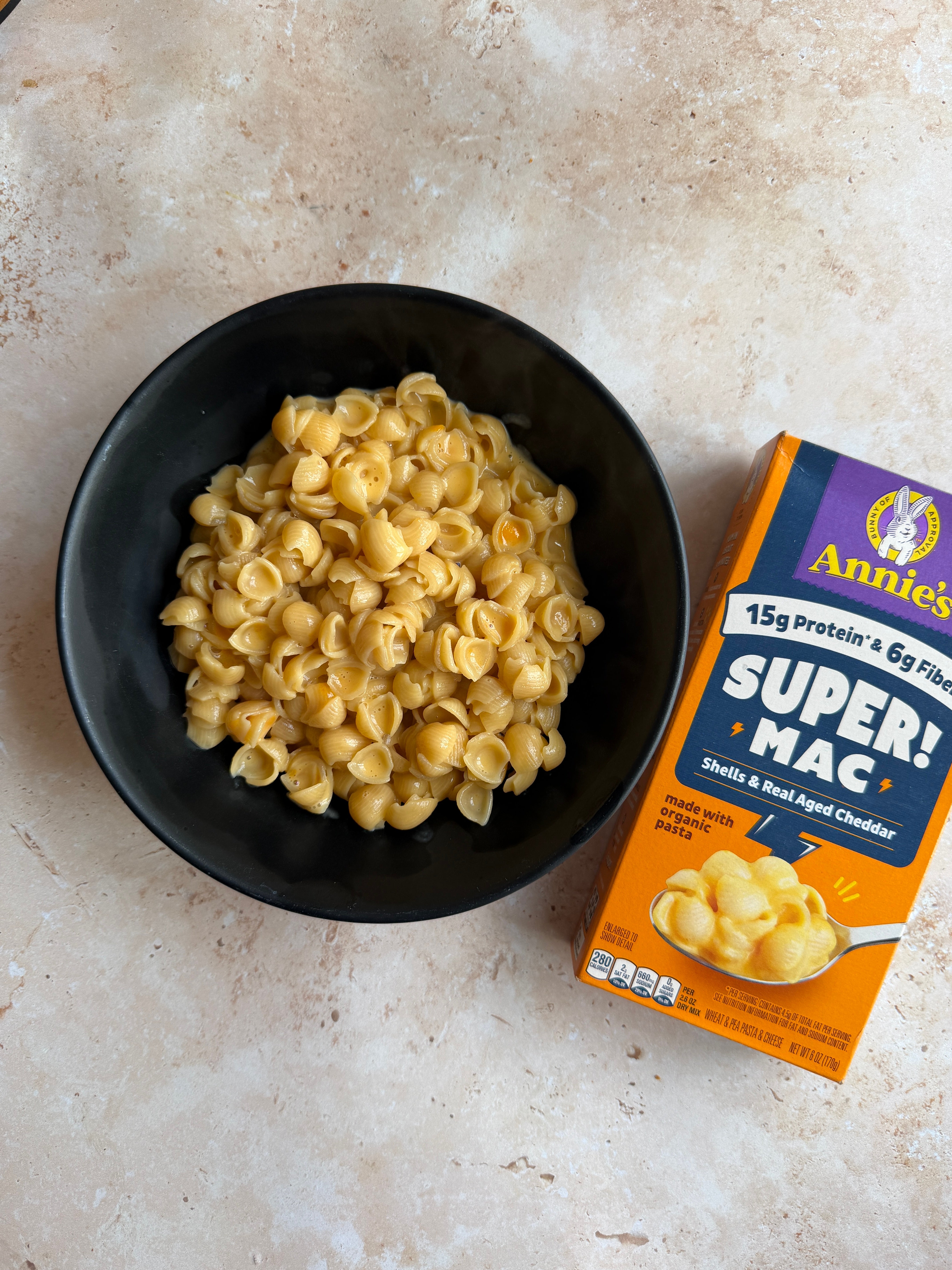 A bowl of shell-shaped macaroni and cheese next to a box labeled "Annie's Super Mac," highlighting protein and fiber content
