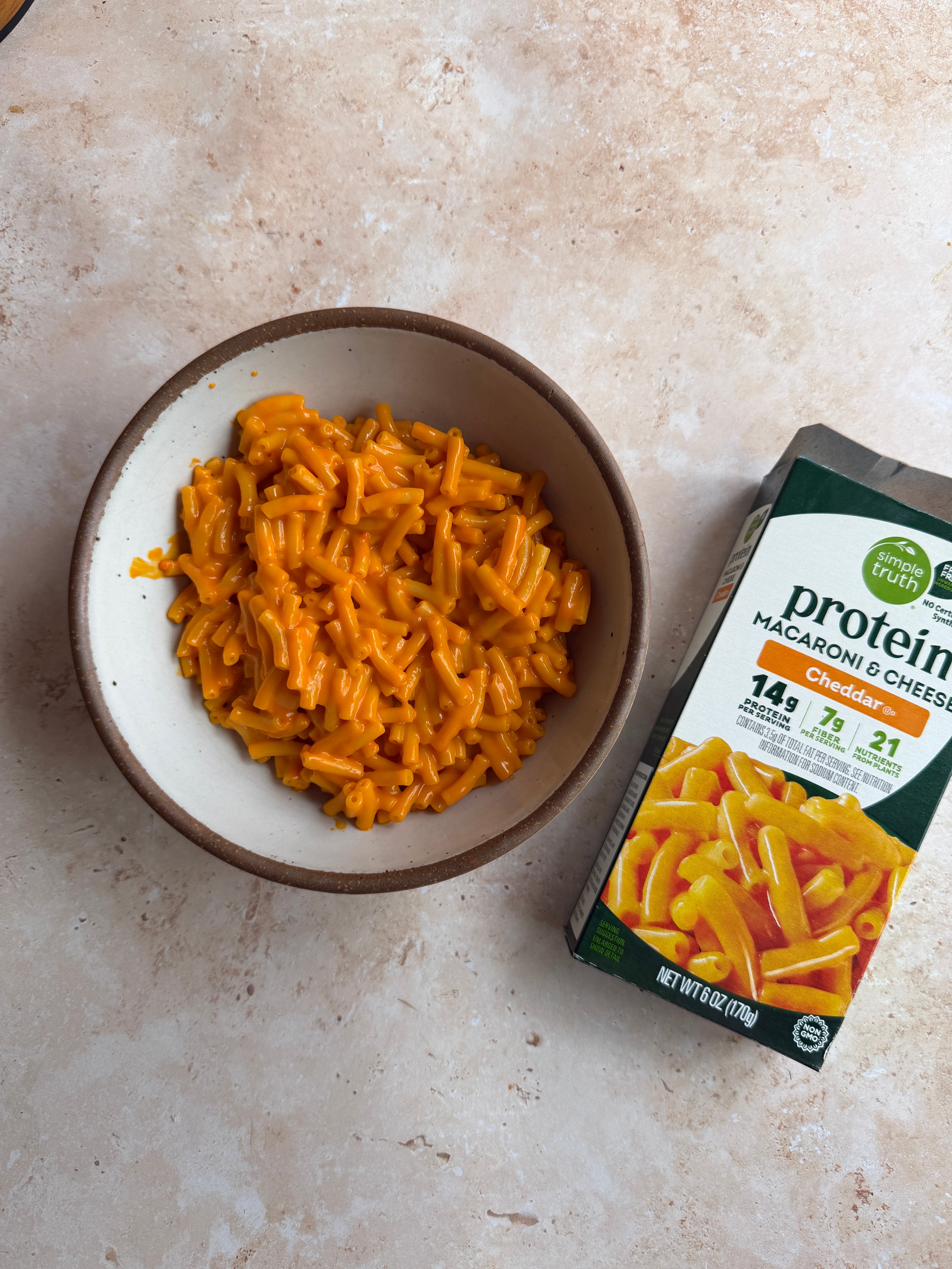 A bowl of cooked macaroni and cheese next to a box labeling it as protein-enriched with cheddar, 14g protein per serving