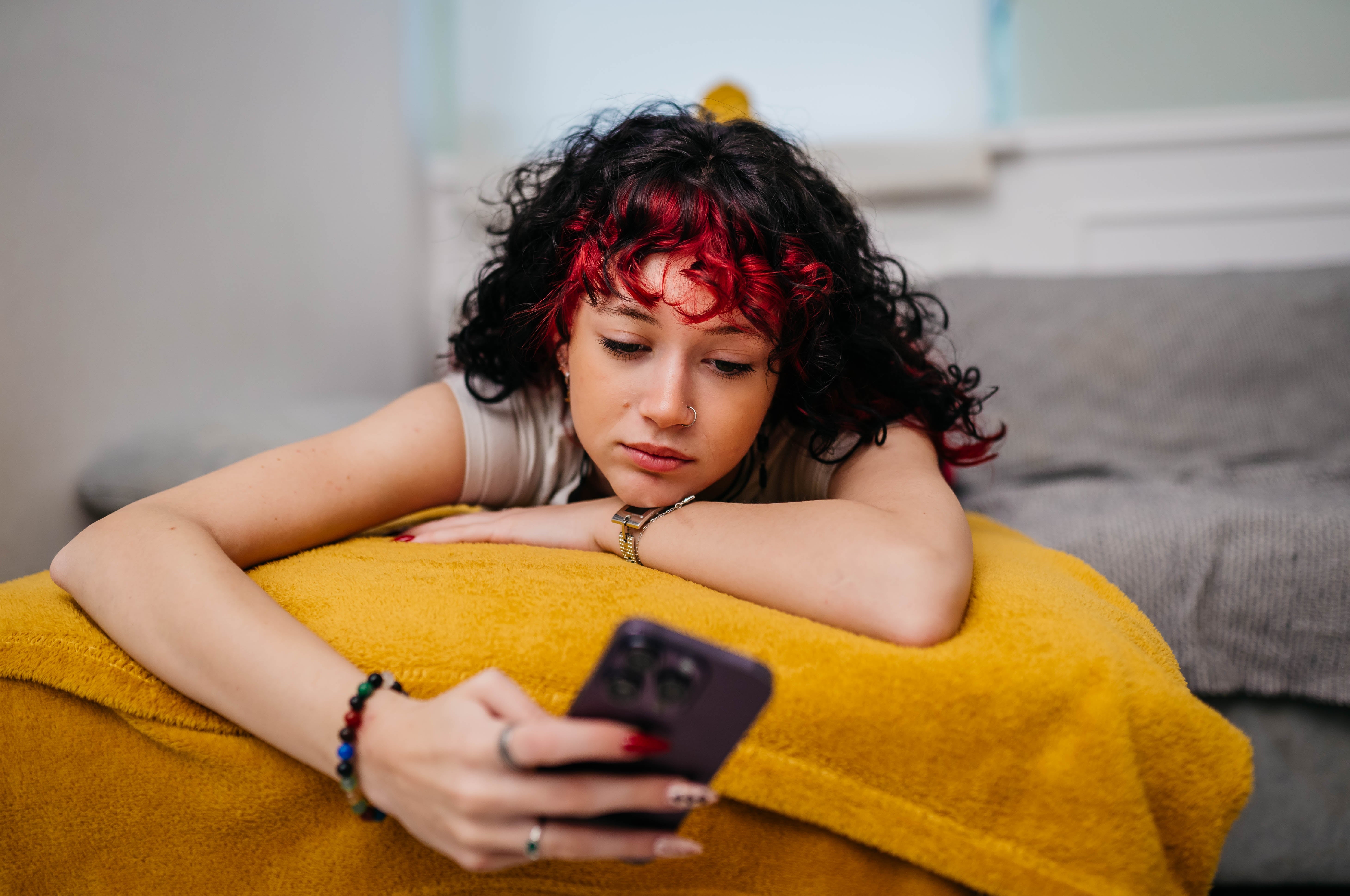 Person with curly hair, styled with vibrant highlights, lying connected  a sofa and looking astatine  a smartphone with a thoughtful expression