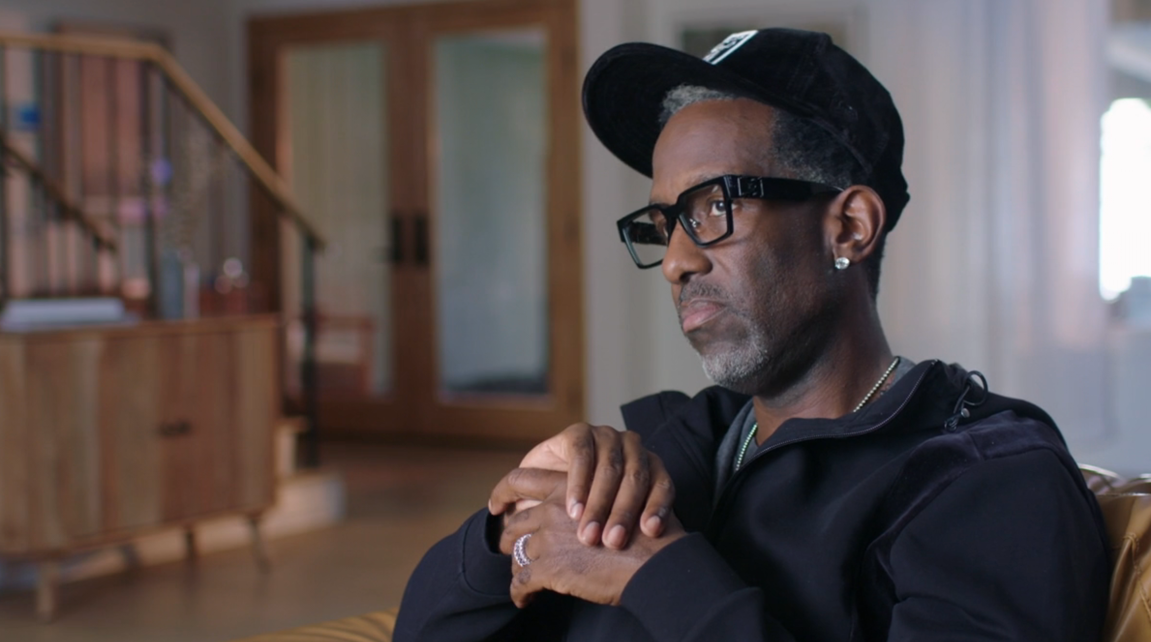 Person wearing glasses and a cap, seated indoors, appears thoughtful with hands clasped. Casual attire with a absorption connected the face