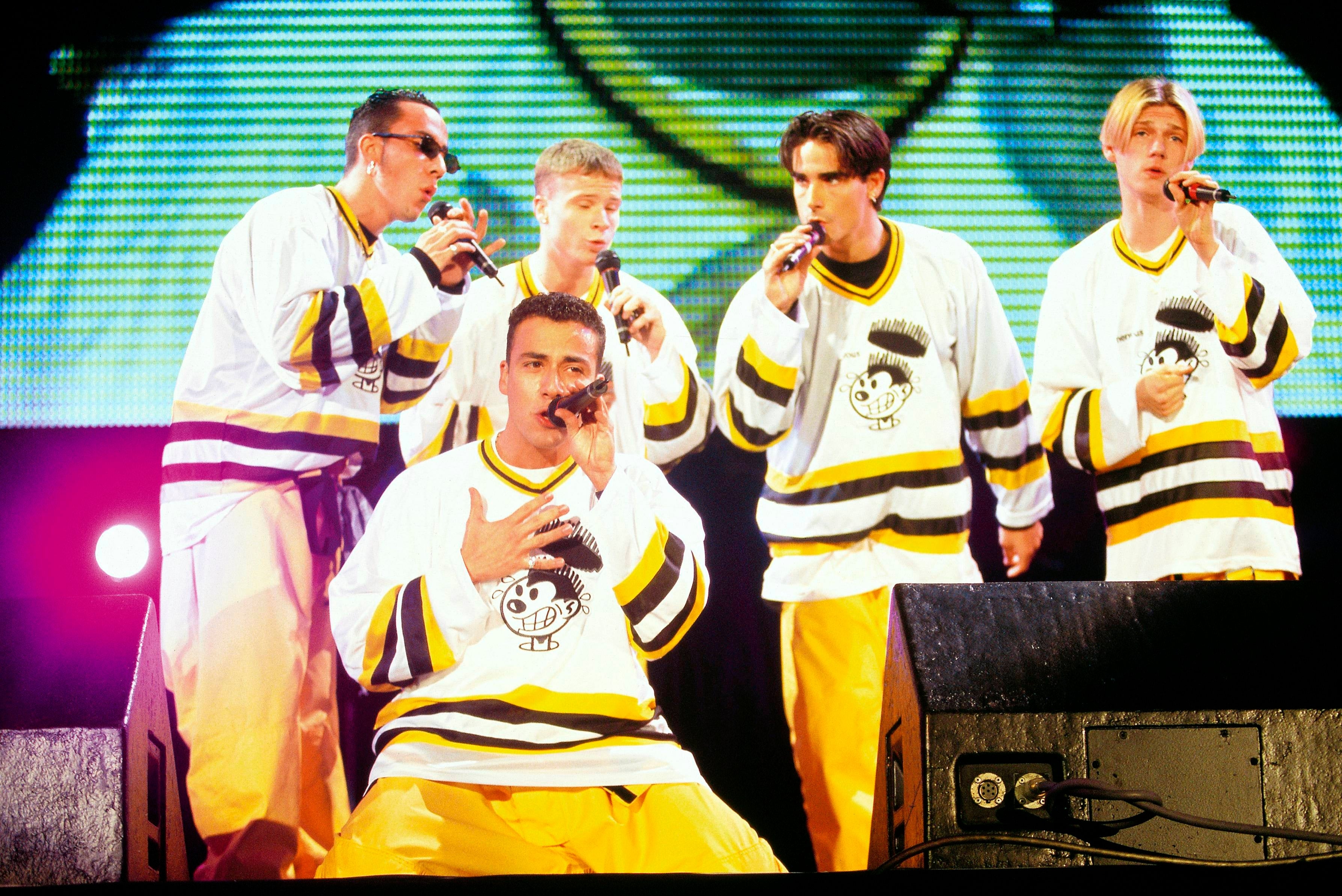 A euphony radical performs connected signifier wearing coordinated jerseys and pants, each holding a microphone, with a ample surface displaying their representation down them