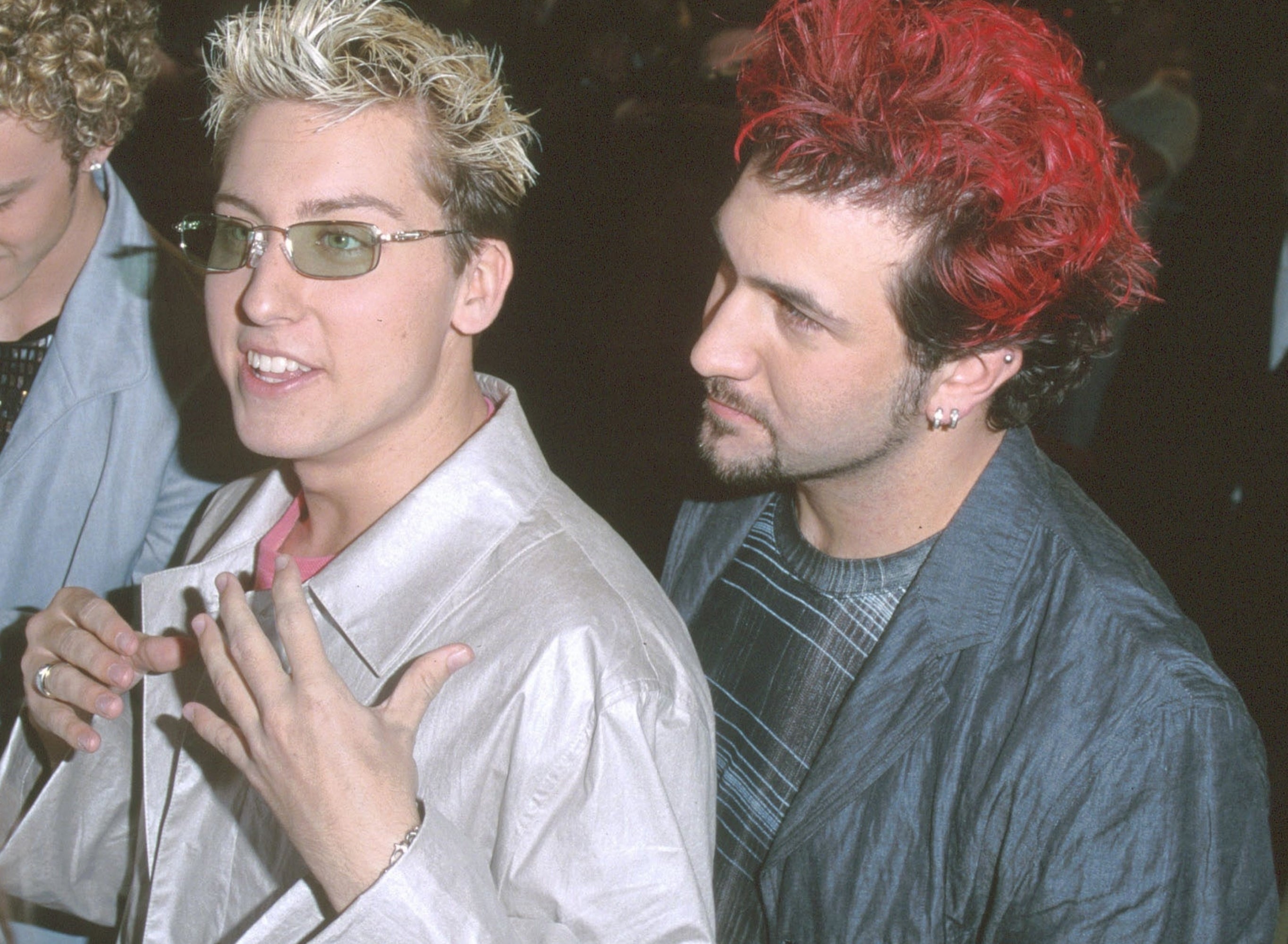 Group of radical interacting with fans, 1 signing autographs. They're wearing stylish, casual attire with unsocial hairstyles