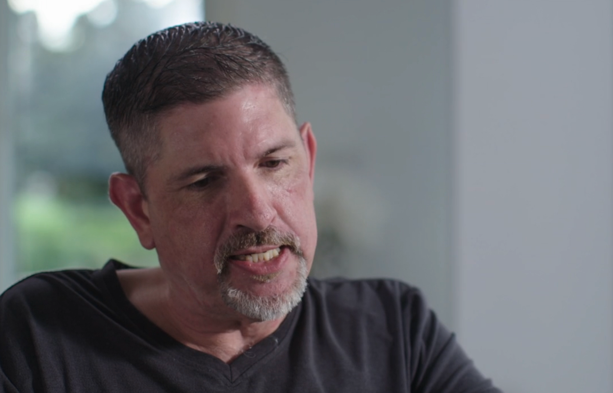 Man with abbreviated hairsbreadth and goatee speaking successful an interrogation setting, wearing a casual acheronian shirt. Background is blurred