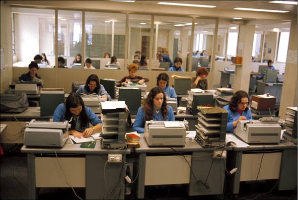People working at desks with typewriters in a busy office setting, focusing on their tasks