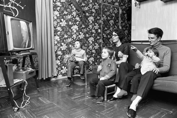 A family of five sits in a living room, engaged and smiling while watching a TV screen