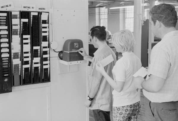 People lined up at a factory punch clock, inserting cards. Two men and one woman wait in line. Time card racks are visible nearby