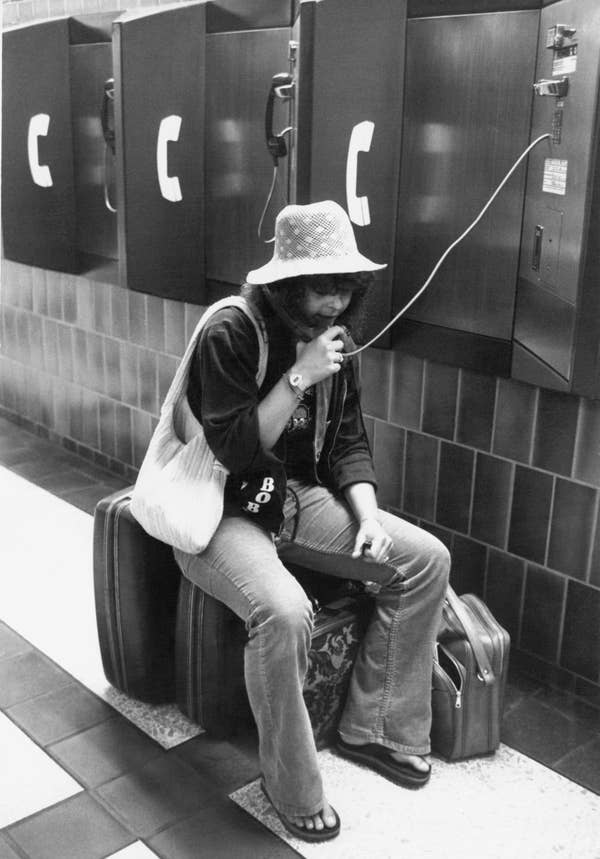Person with hat using a payphone, sitting on luggage. Retro setting with wall-mounted phones