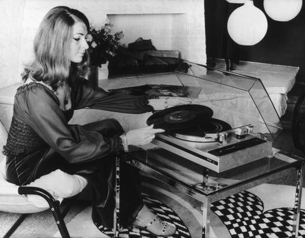 Woman in elegant 1970s attire placing a vinyl record on a turntable in a stylish living room