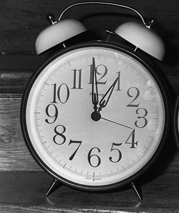 A classic analog alarm clock shows 10:09. It has two bells on top and is placed on a wooden surface