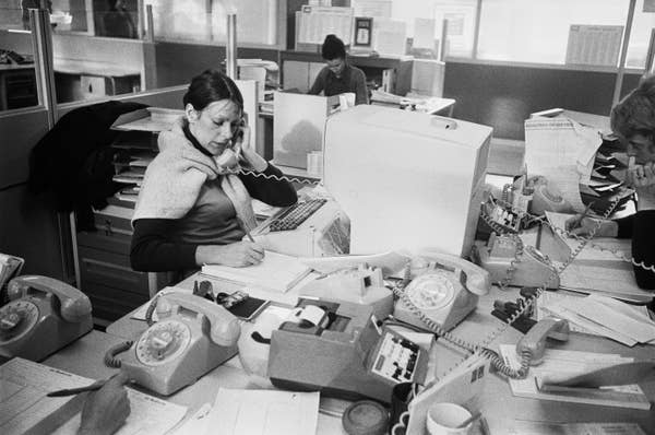 A vintage office scene with three people working at desks filled with papers, rotary phones, and an early computer