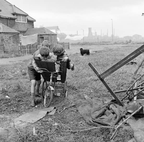 Two boys play with a homemade go-kart in a grassy, industrial area. One boy rides while the other pushes, with factory smokestacks in the background