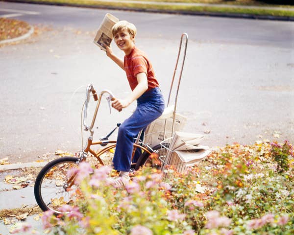 Teen delivering newspapers on a bicycle, smiling, with a basket full of newspapers in a suburban setting