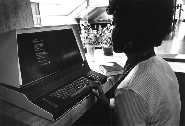 Person using a vintage computer with text on the screen, sitting at a desk in a well-lit room with books and potted plants