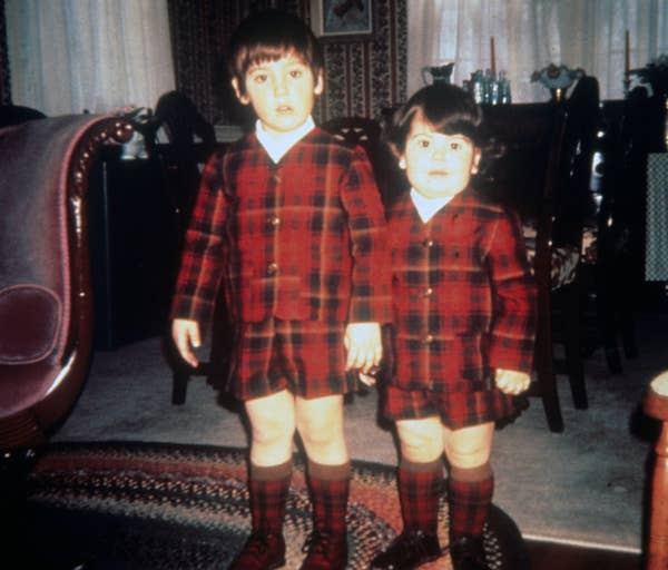 Two young children wearing matching plaid outfits, standing in a room with a patterned rug and furniture in the background