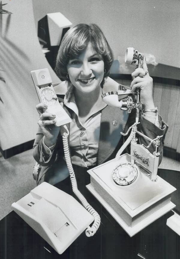 Person smiling, holding a vintage telephone and handset. The person is wearing a blazer and seated at a table with two telephones in front