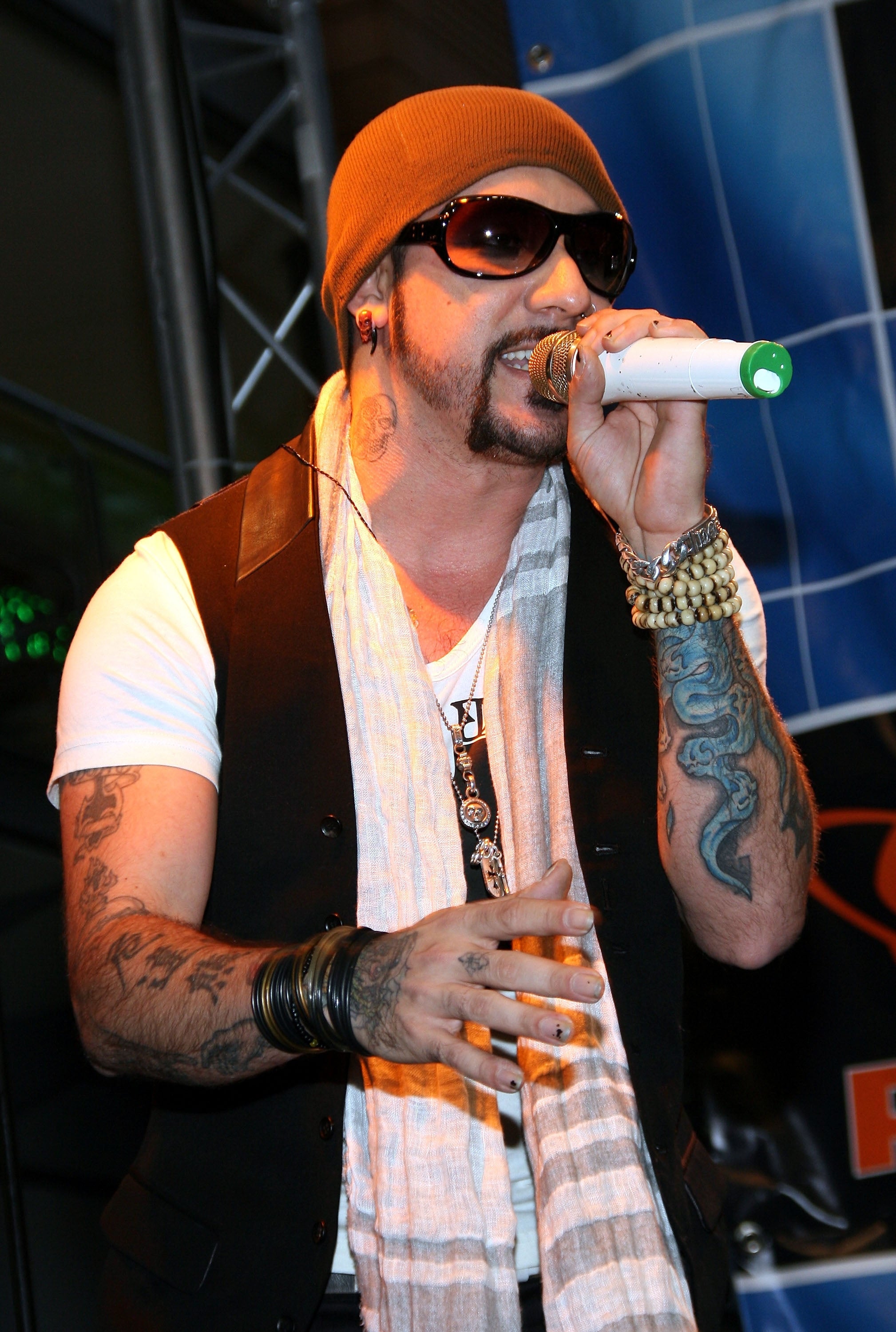 A idiosyncratic with tattoos and sunglasses performs connected stage, wearing a beanie, a vest implicit a T-shirt, and aggregate bracelets