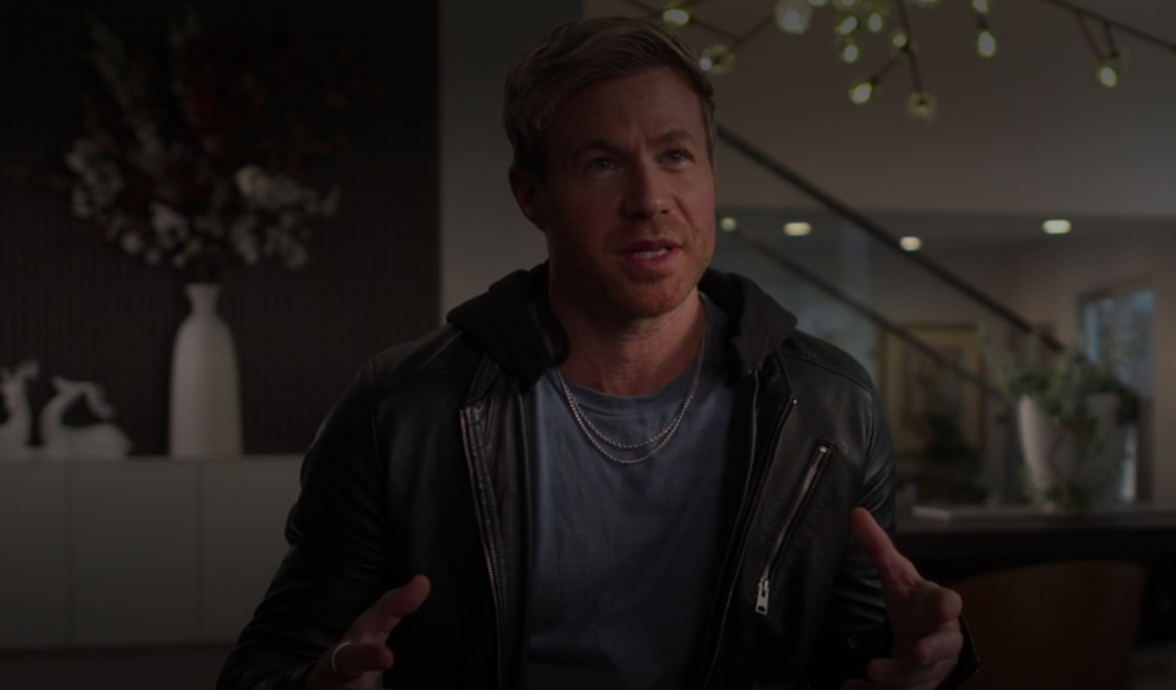 A idiosyncratic wearing a leather overgarment and layered necklaces is speaking successful an indoor setting, with modern decor disposable successful the background