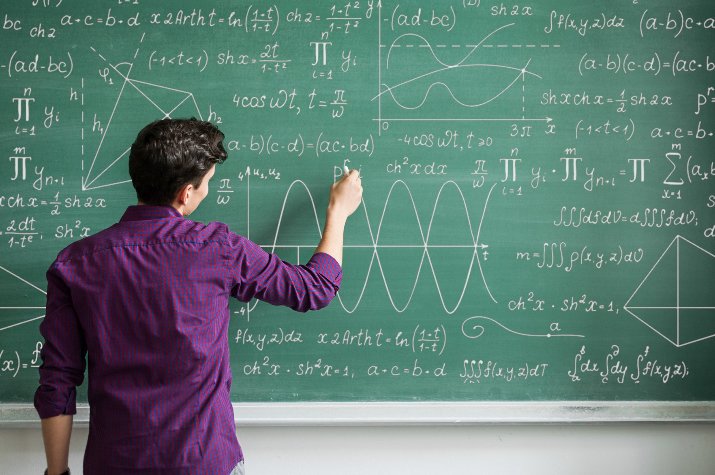 Person writing complex mathematical equations and sketches on a large chalkboard filled with various symbols and formulas