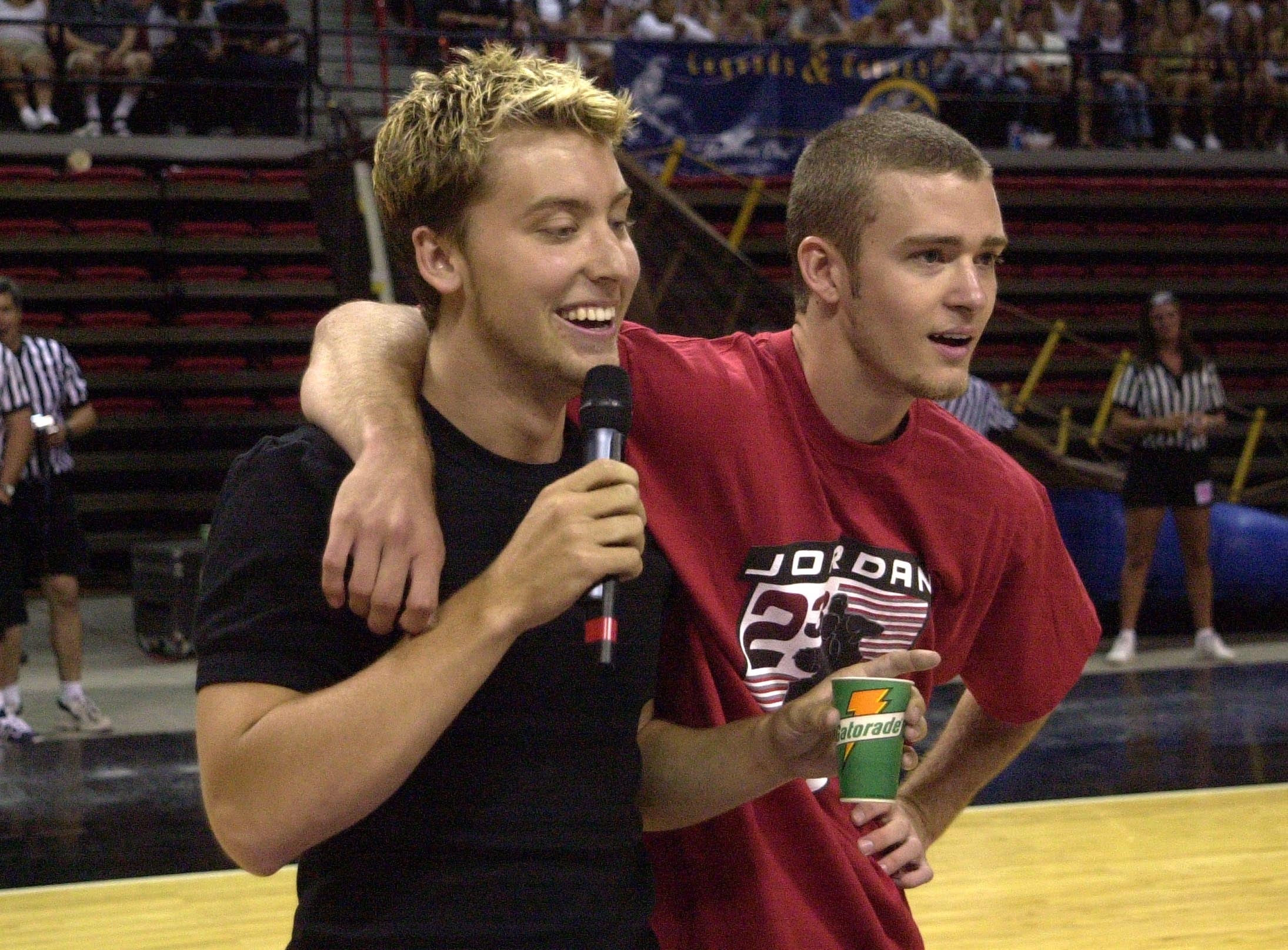 Two individuals connected a hoops court; 1 is holding a microphone with an limb astir the other, who is successful a casual t-shirt holding a drink