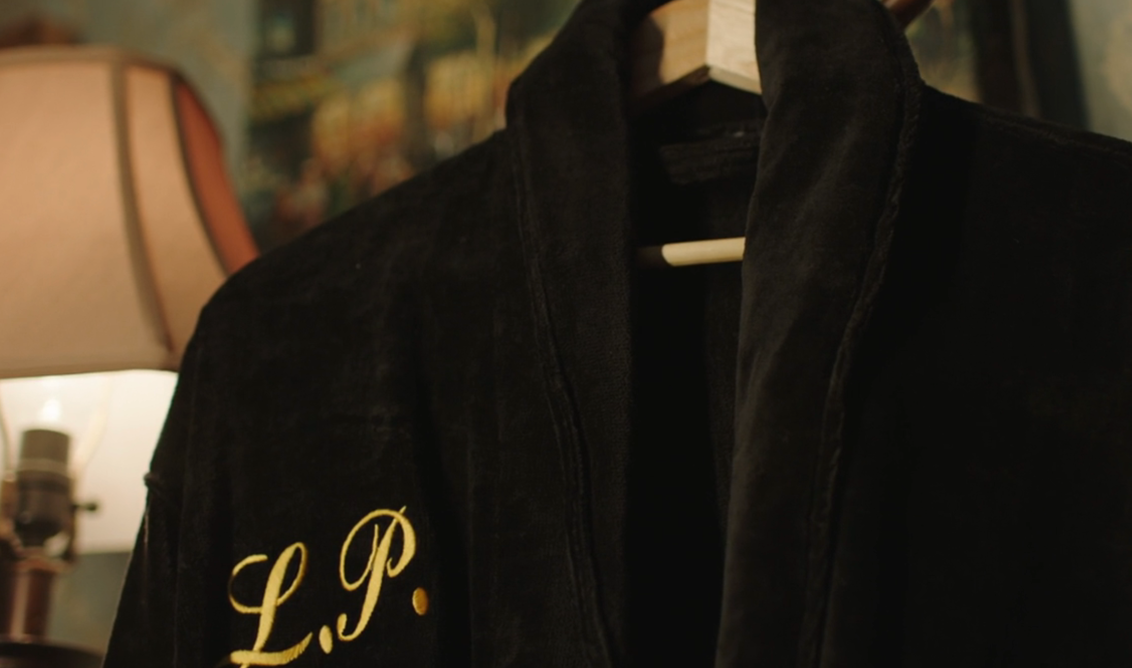 Monogrammed robe with "L.P." hangs connected a woody hanger. A lamp and blurred inheritance are visible