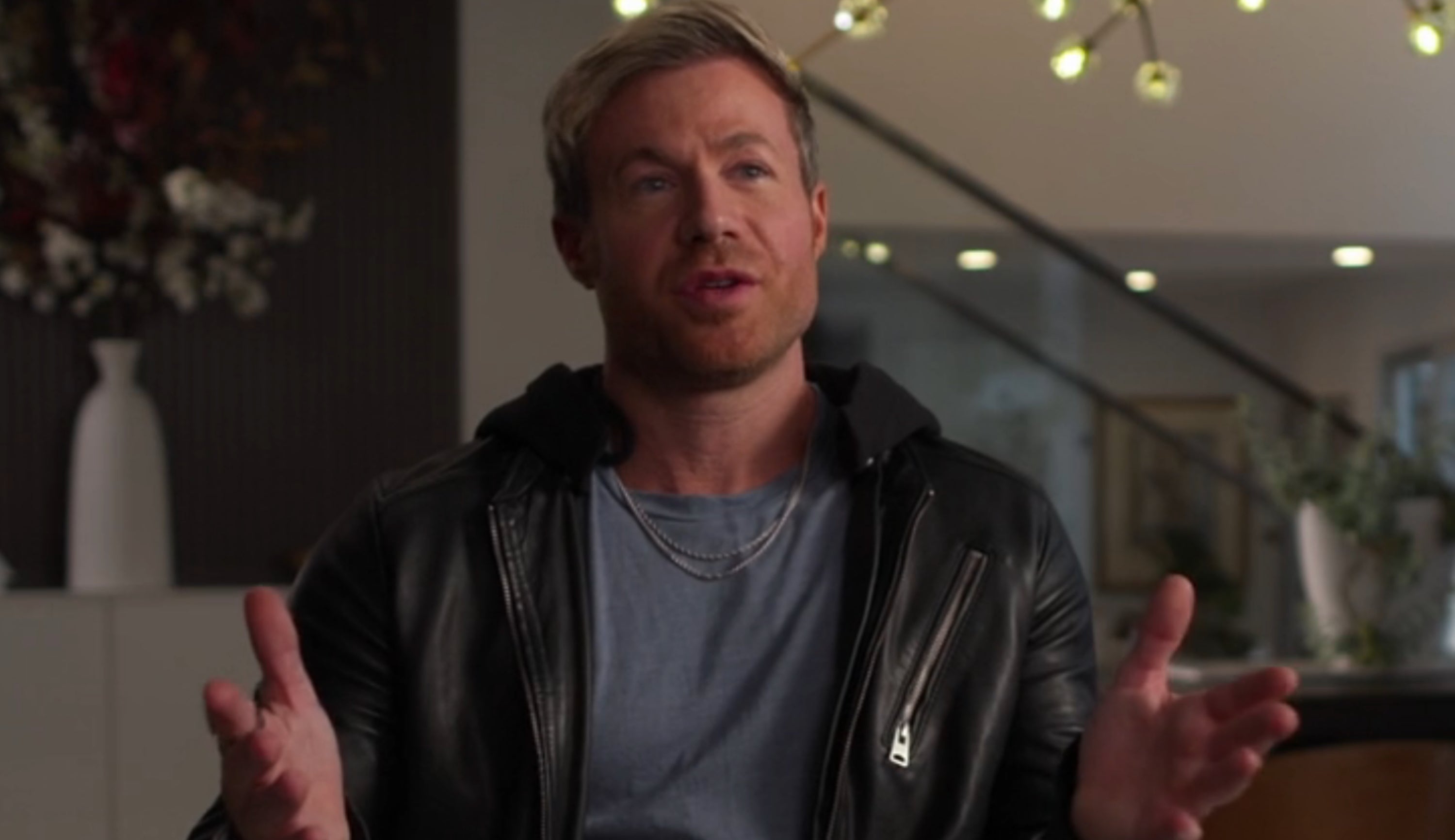 A idiosyncratic is seated indoors, gesturing portion talking. They are wearing a casual leather jacket. Text identifies them arsenic an artist