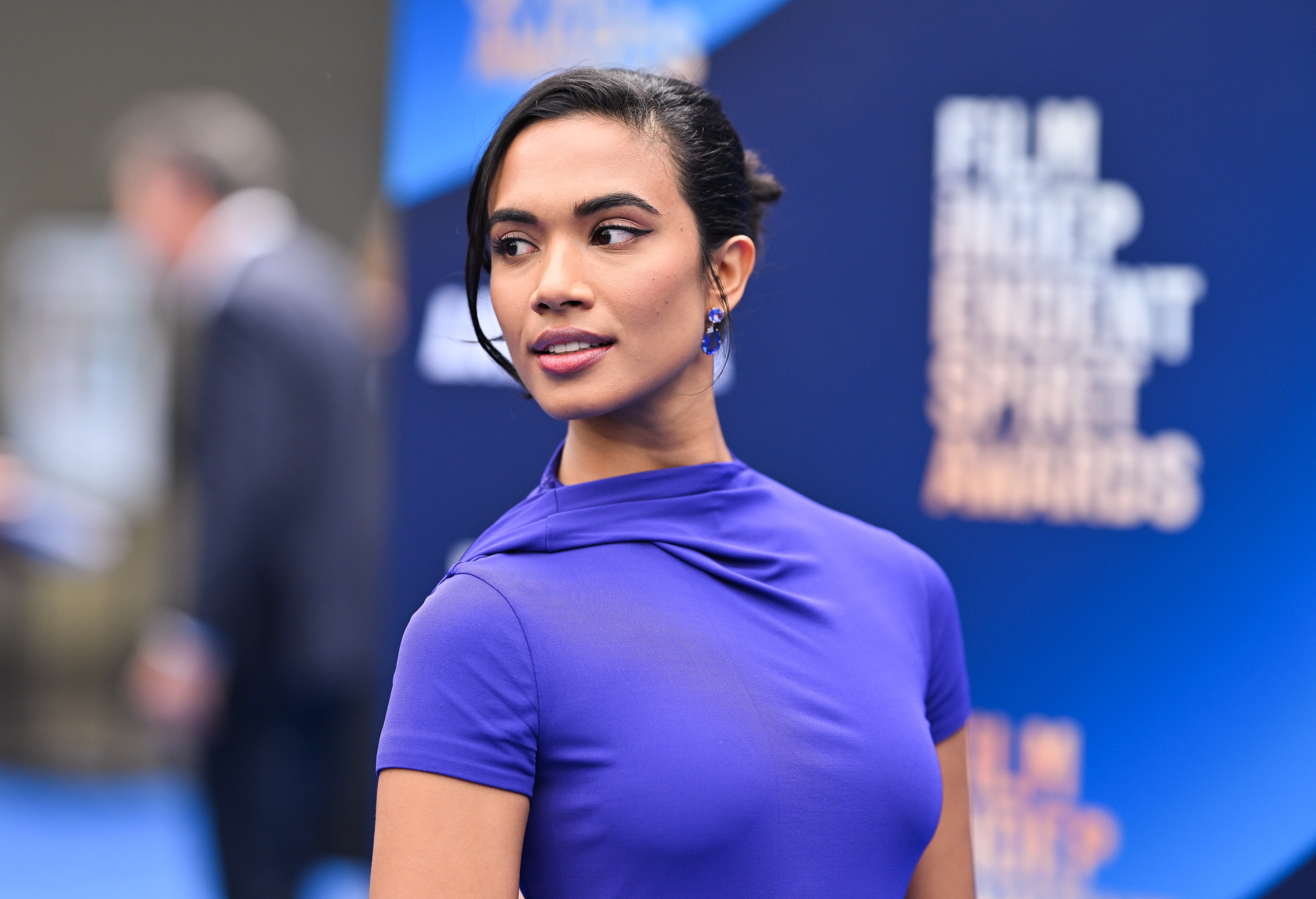 Person connected  reddish  carpet successful  elegant, fitted formal  with precocious   neckline. Hair styled up, wearing earrings. Blue-themed inheritance  visible