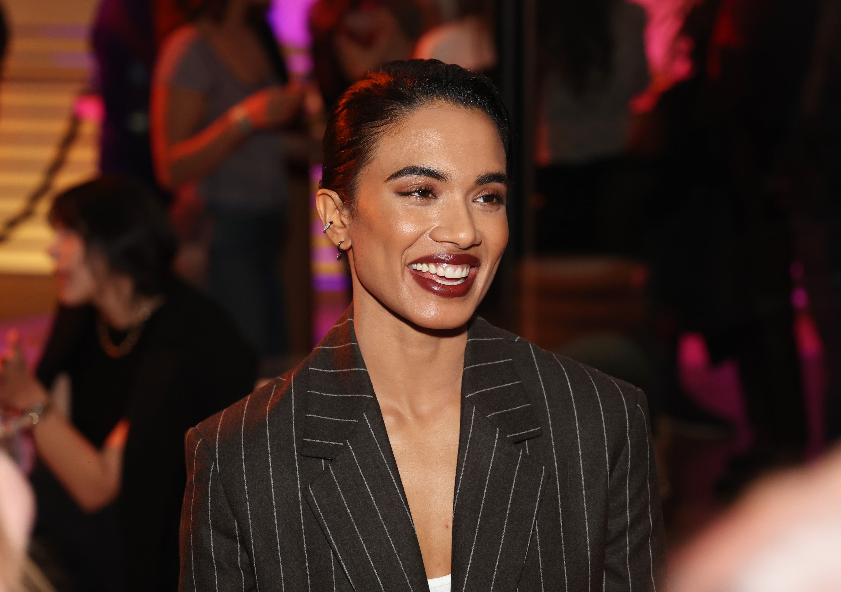 Person wearing a pinstripe suit   smiles astatine  an indoor event. Socializing radical   and colorful lights are successful  the background