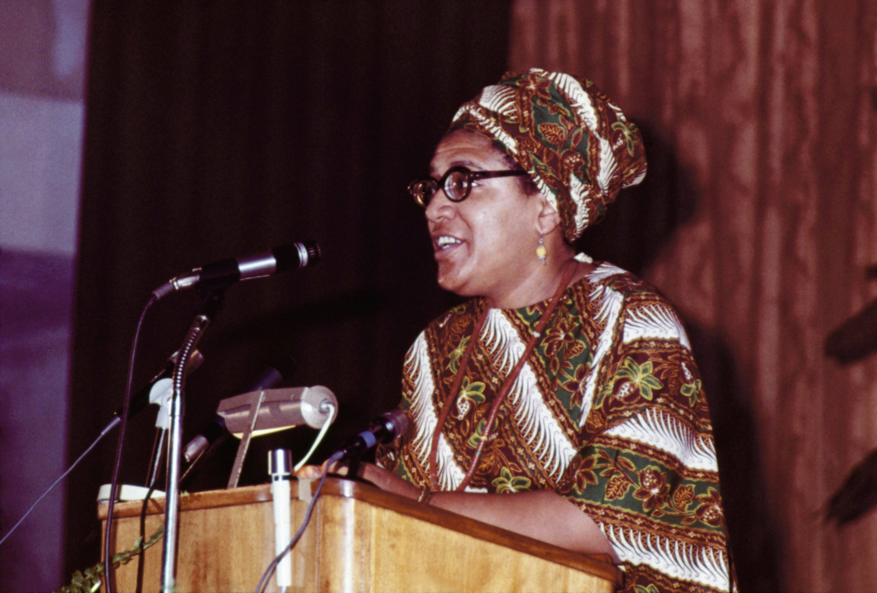 Person astatine  a podium speaking into microphones, wearing patterned accepted   attire and a matching headwrap