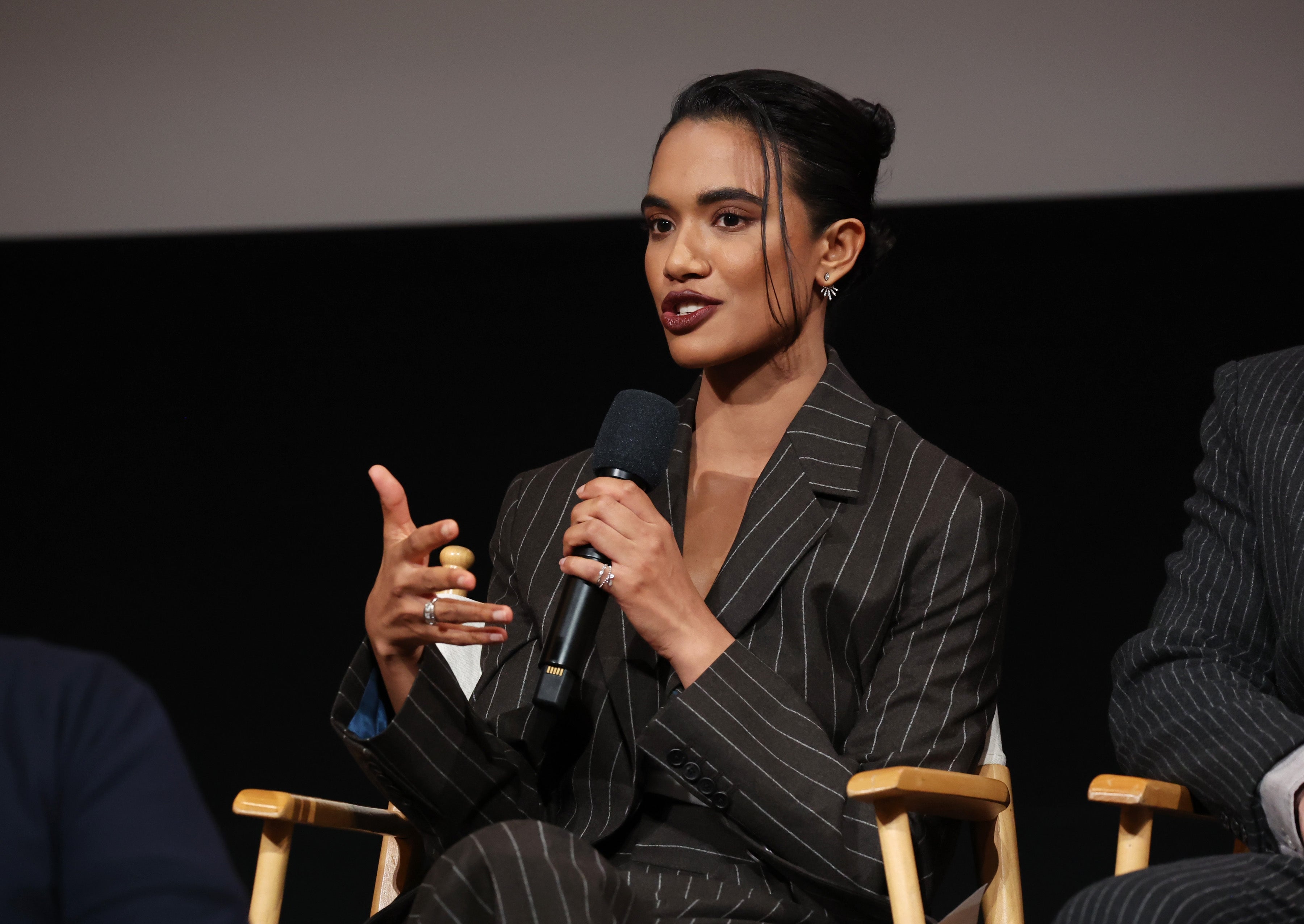 Person speaking connected  stage, wearing a pinstripe suit, holding a microphone, gesturing with 1  hand