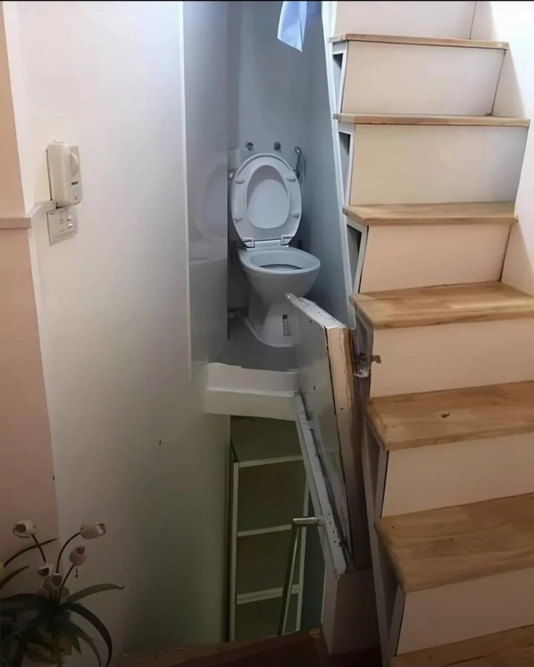 Toilet awkwardly positioned nether  stairs, partially blocking a constrictive  passageway with constricted  accessibility