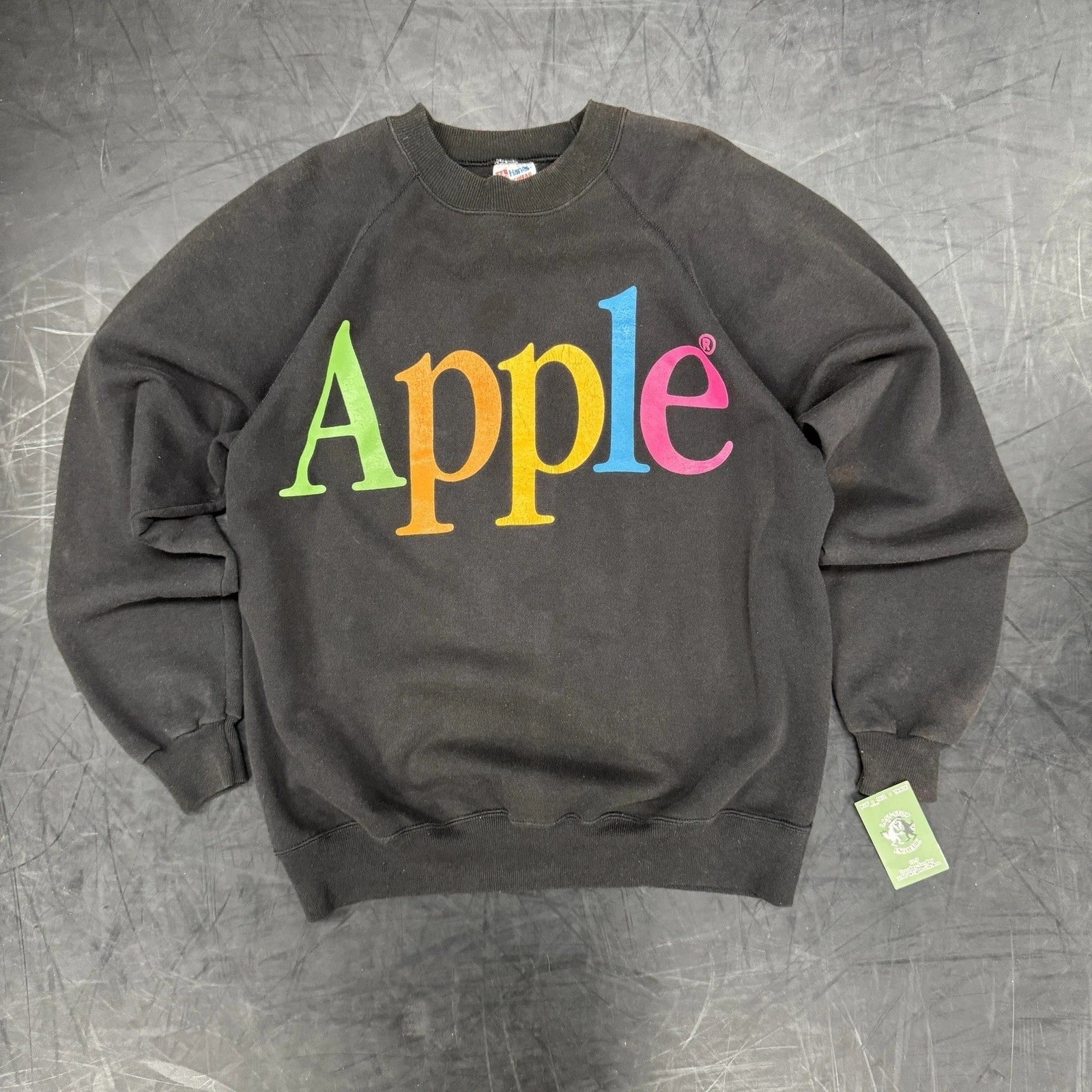 Black sweatshirt featuring a retro Apple logo with multicolored letters displayed connected  a grey  surface