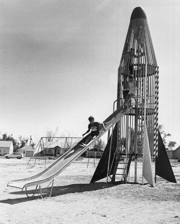 Children playing on a retro rocket-themed playground slide in an open park area