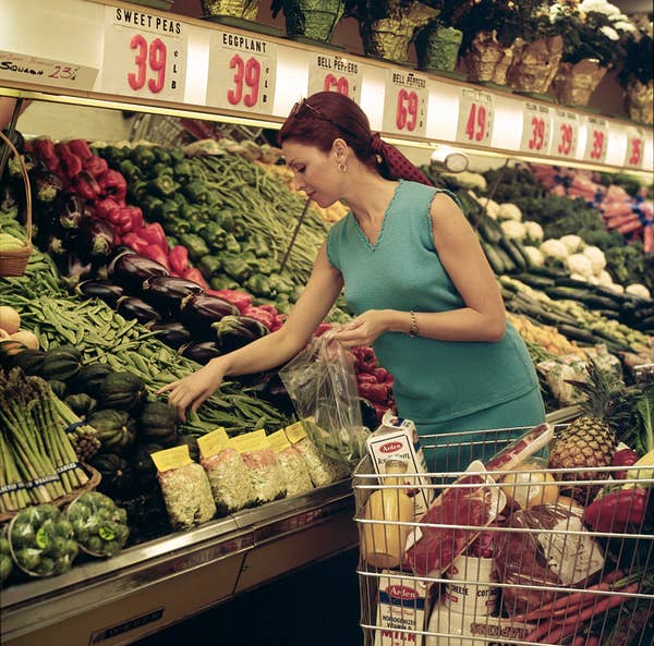 A woman in a sleeveless top shops for fresh produce, picking vegetables, with a full cart nearby in a grocery store