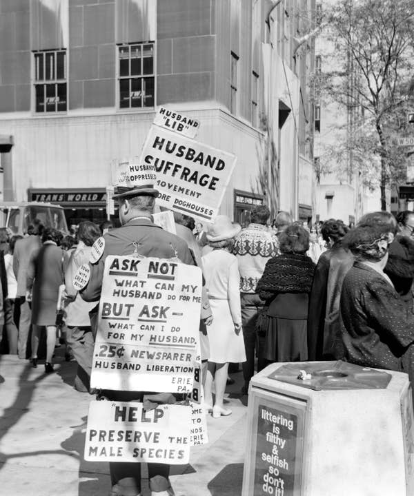 People in a 1960s protest holding satirical signs advocating for "husband suffrage" and "husband liberation" in a city street
