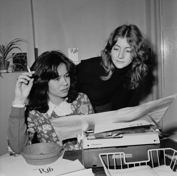Two women at a desk with a typewriter appear focused on reading a document. One is seated with a cigarette, both look engaged in their work