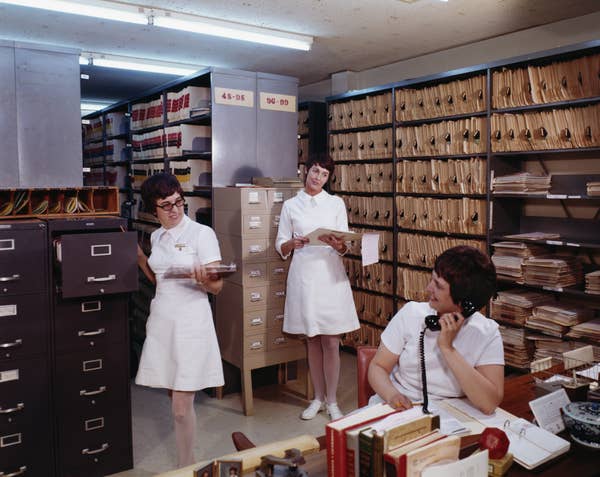 Three women in matching dresses work in an organized file room, each handling documents or on a phone. The room has shelves filled with files