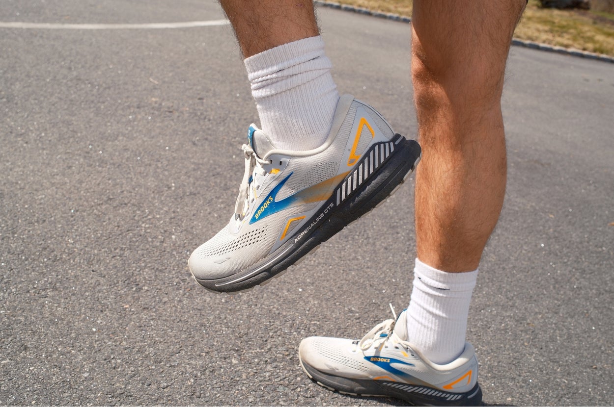 Person wearing athletic sneakers and white socks while standing mid-step on a paved surface. Focus on the shoe design, suited for active lifestyle