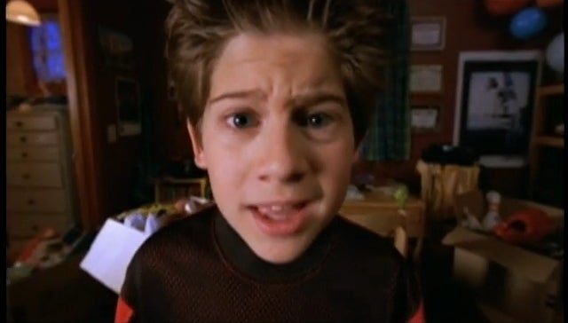 A young person with spiky hair looks directly at the camera with a surprised expression, in a cluttered room
