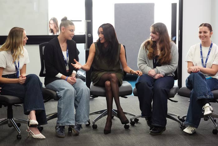 A pistillate   successful  a stylish outfit interacts animatedly with 4  young women seated successful  an bureau   setting