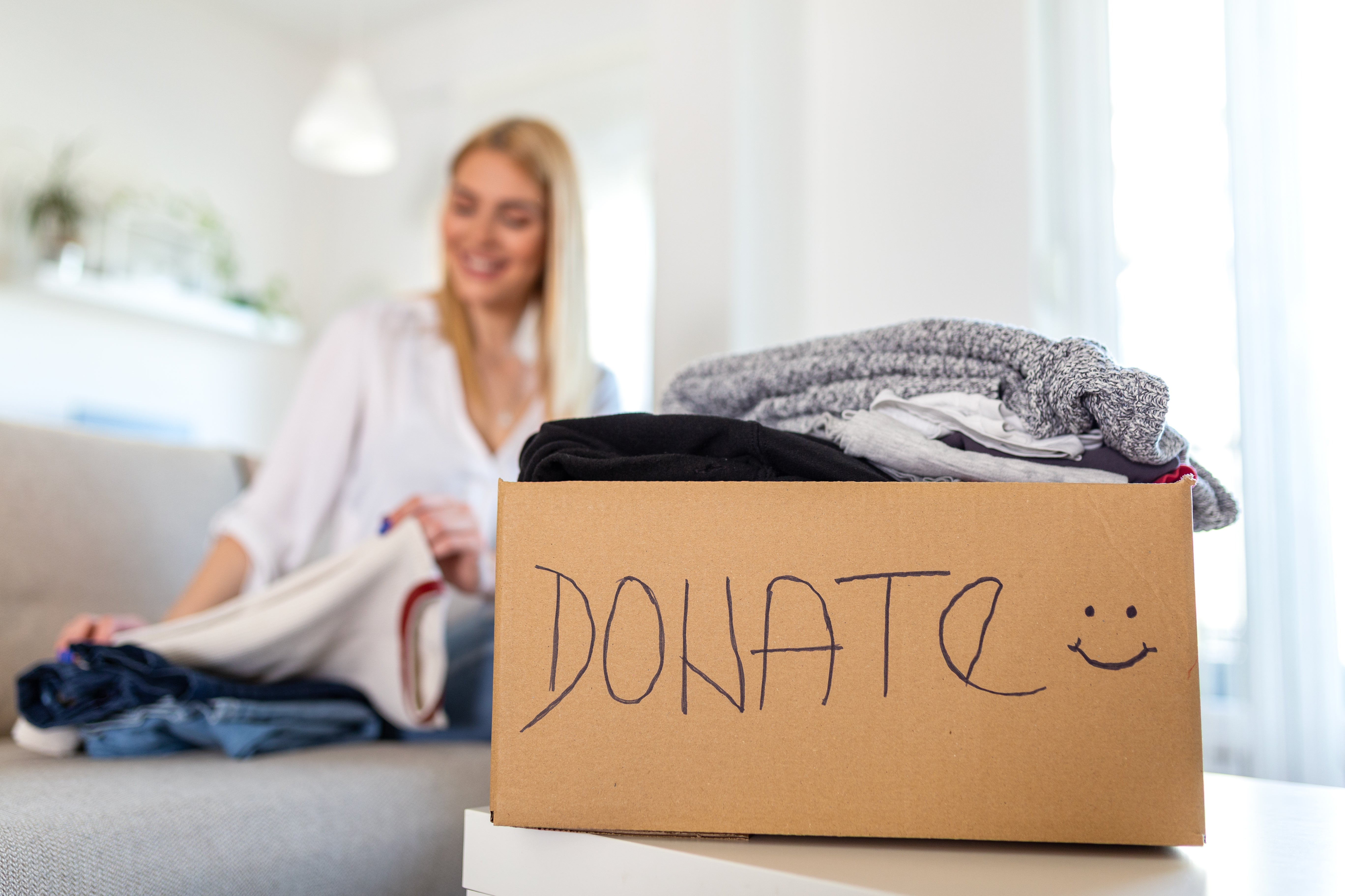 Woman organizing clothes with a cardboard box labeled "Donate" filled with folded garments in the foreground