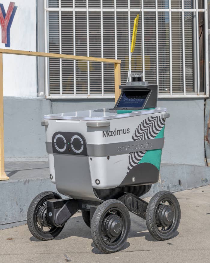 Delivery robot on wheels with a "Maximus" label, parked on a sidewalk, seen in an urban environment