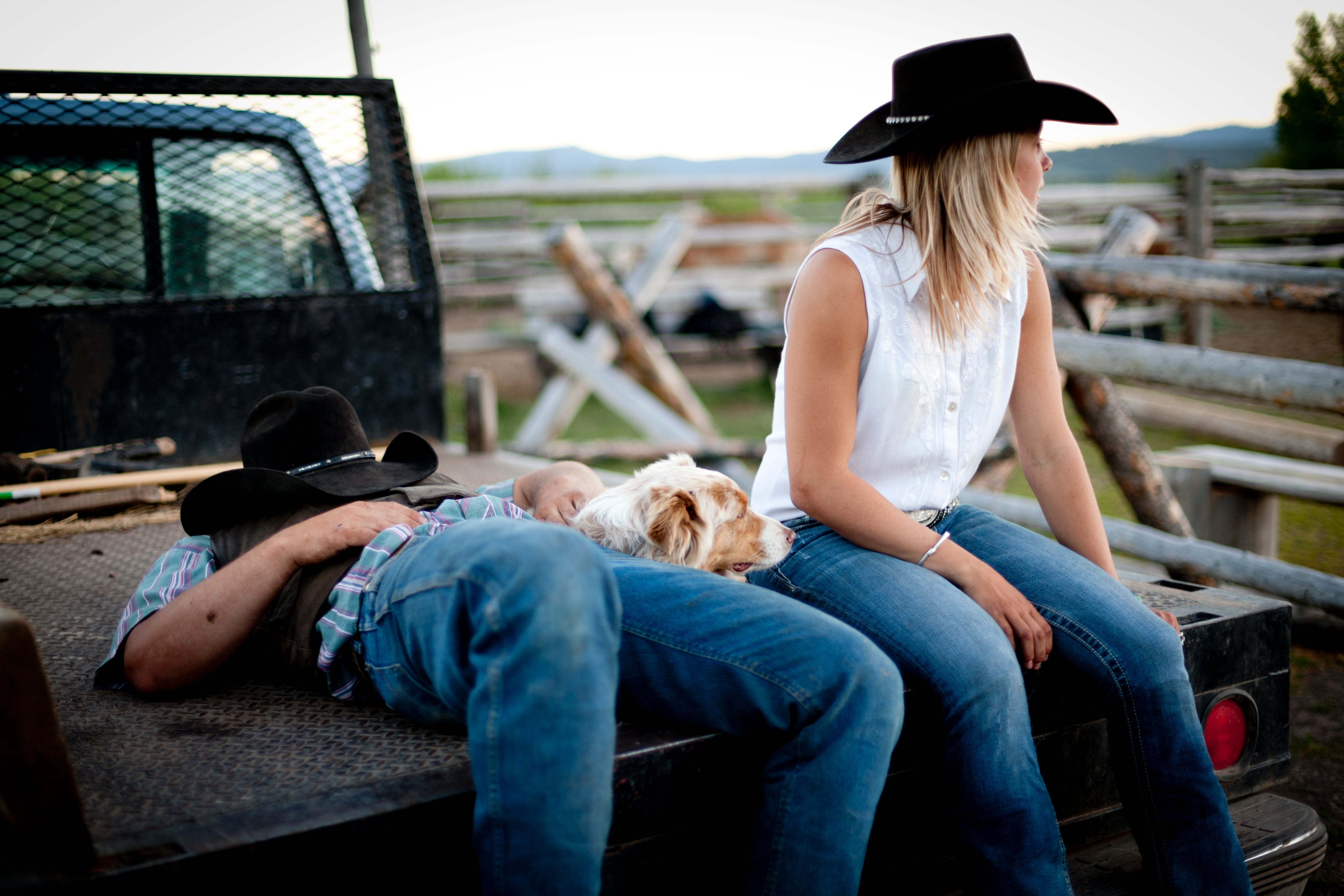 Two people in cowboy hats relax on a truck bed, one resting with a dog. The scene suggests a casual, intimate moment in a rural setting