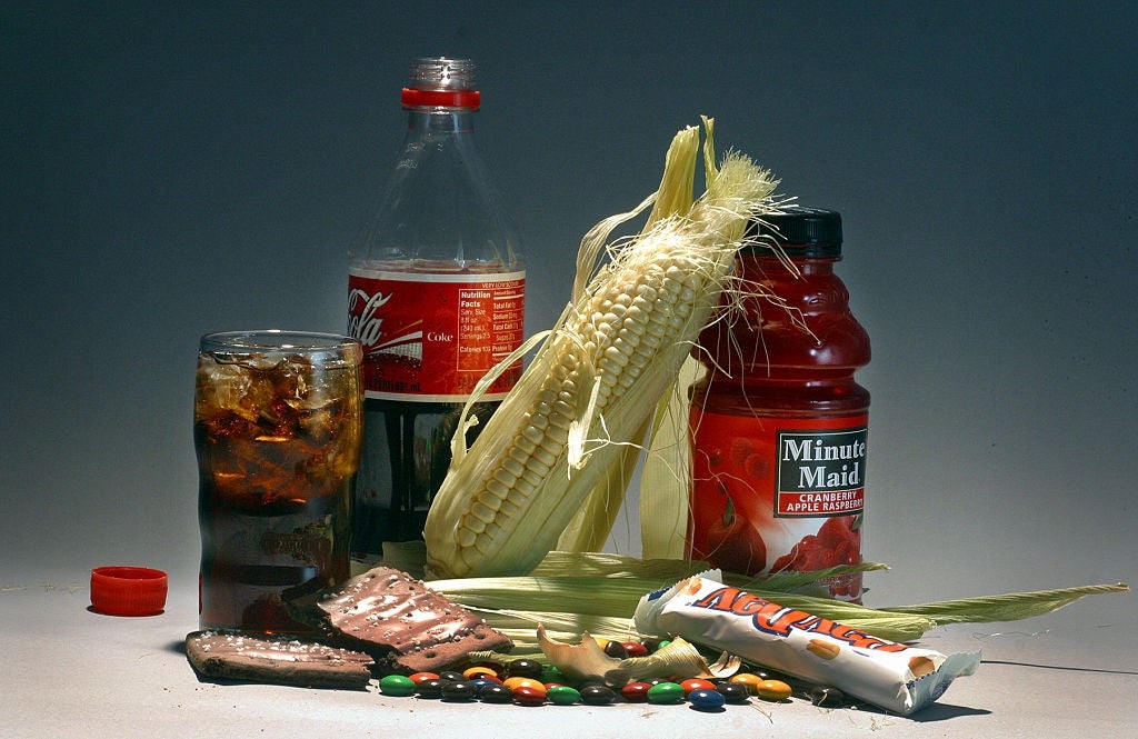 An assortment of sugar-rich foods and drinks, including soda, corn, juice, cocoa  bar, and candies, displayed connected  a surface
