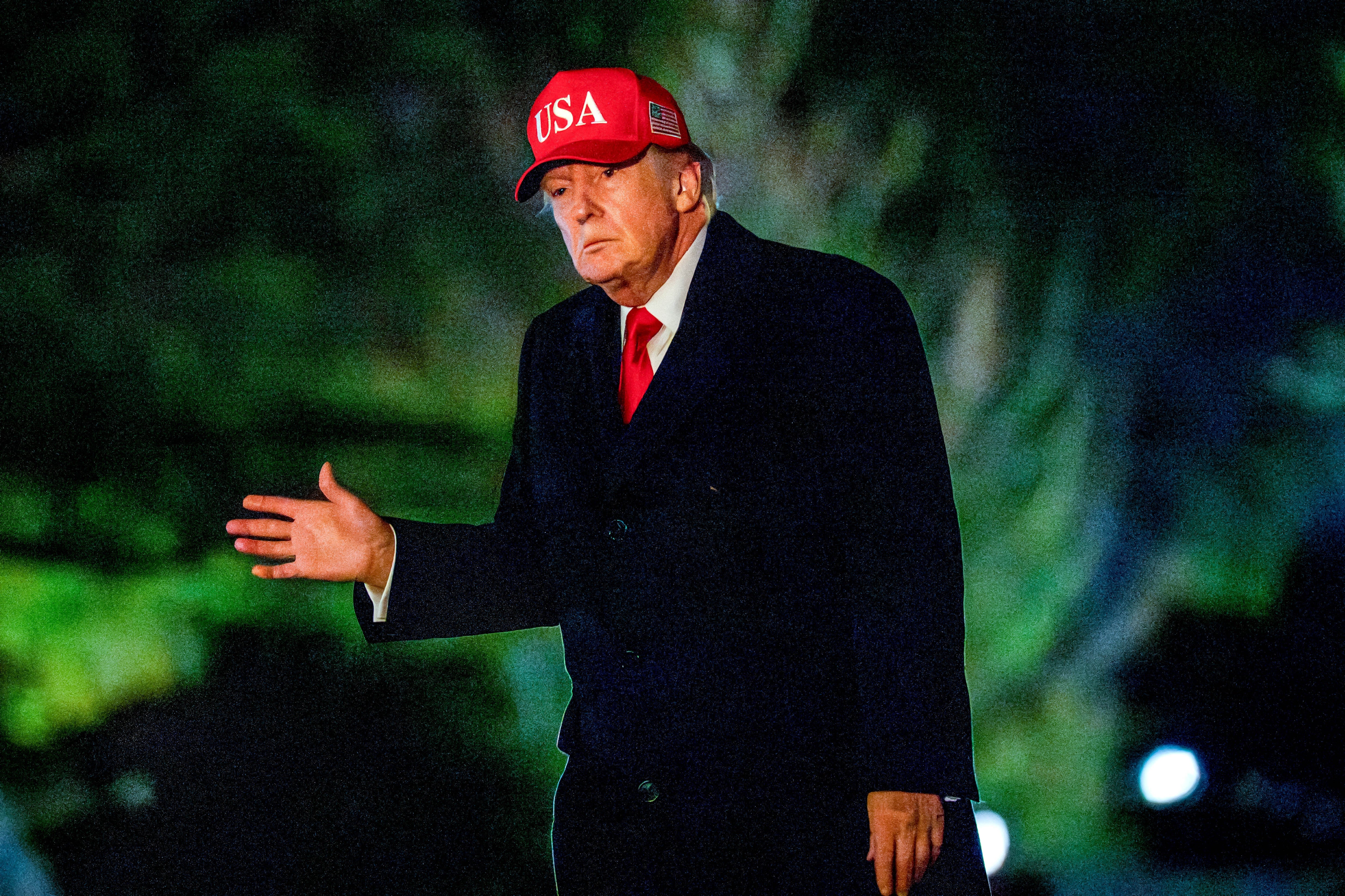 Man successful  a acheronian  suit   and reddish  necktie  wears a chapeau  with "USA" and gestures with 1  hand, lasting  outdoors astatine  night