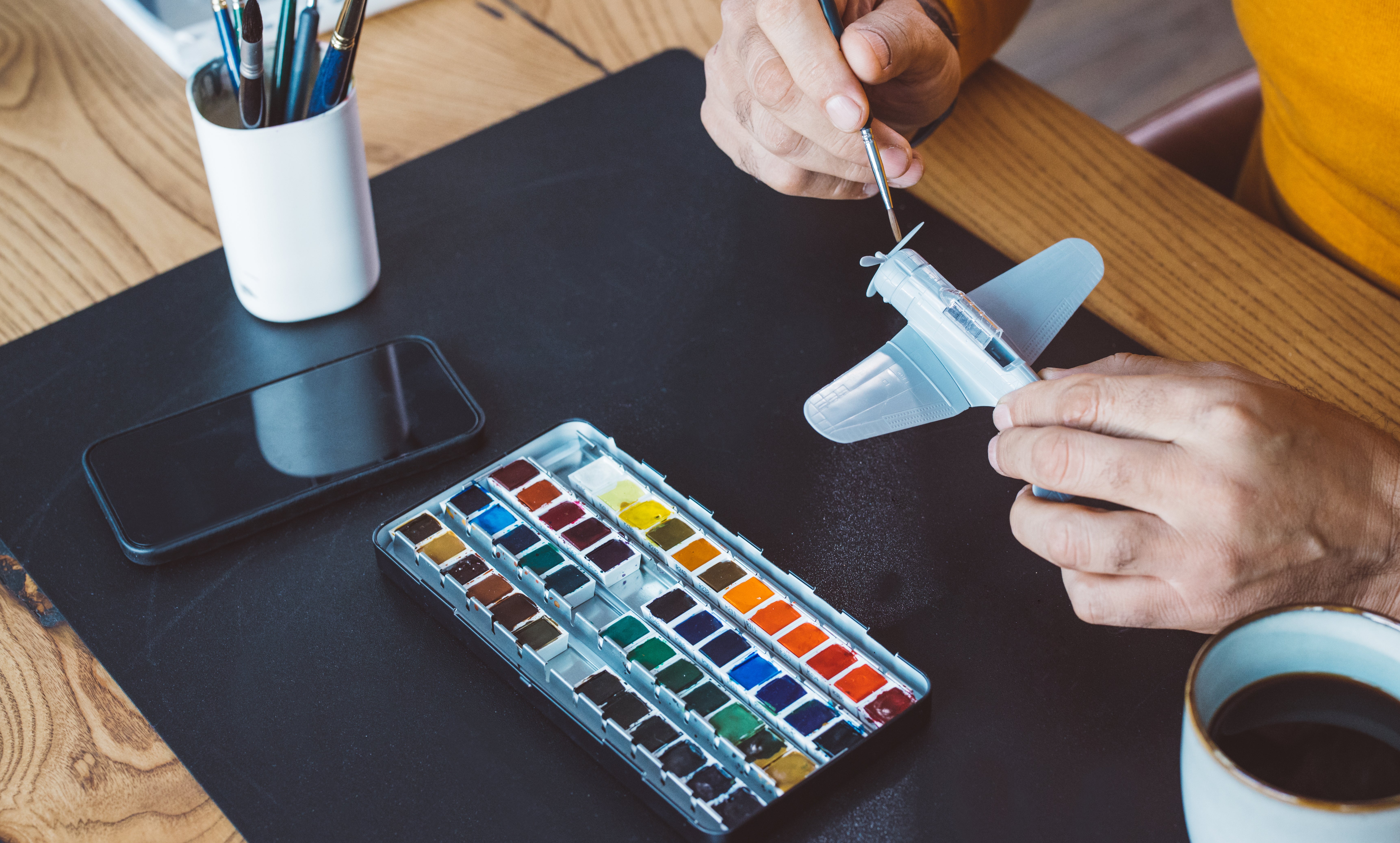 Person coating  a tiny  exemplary  airplane, surrounded by watercolor paints, brushes, phone, and a java  cupful  connected  a woody  table