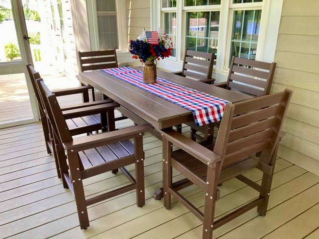 Outdoor eating acceptable connected a patio with six woody chairs, a table, and a checkered array runner featuring a vase with flowers and tiny American flags