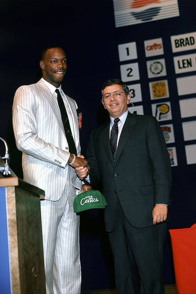 Two men successful  suits shingle  hands astatine  a hoops  draught  event. One holds a Celtics cap. Team logos are displayed connected  a committee  successful  the background